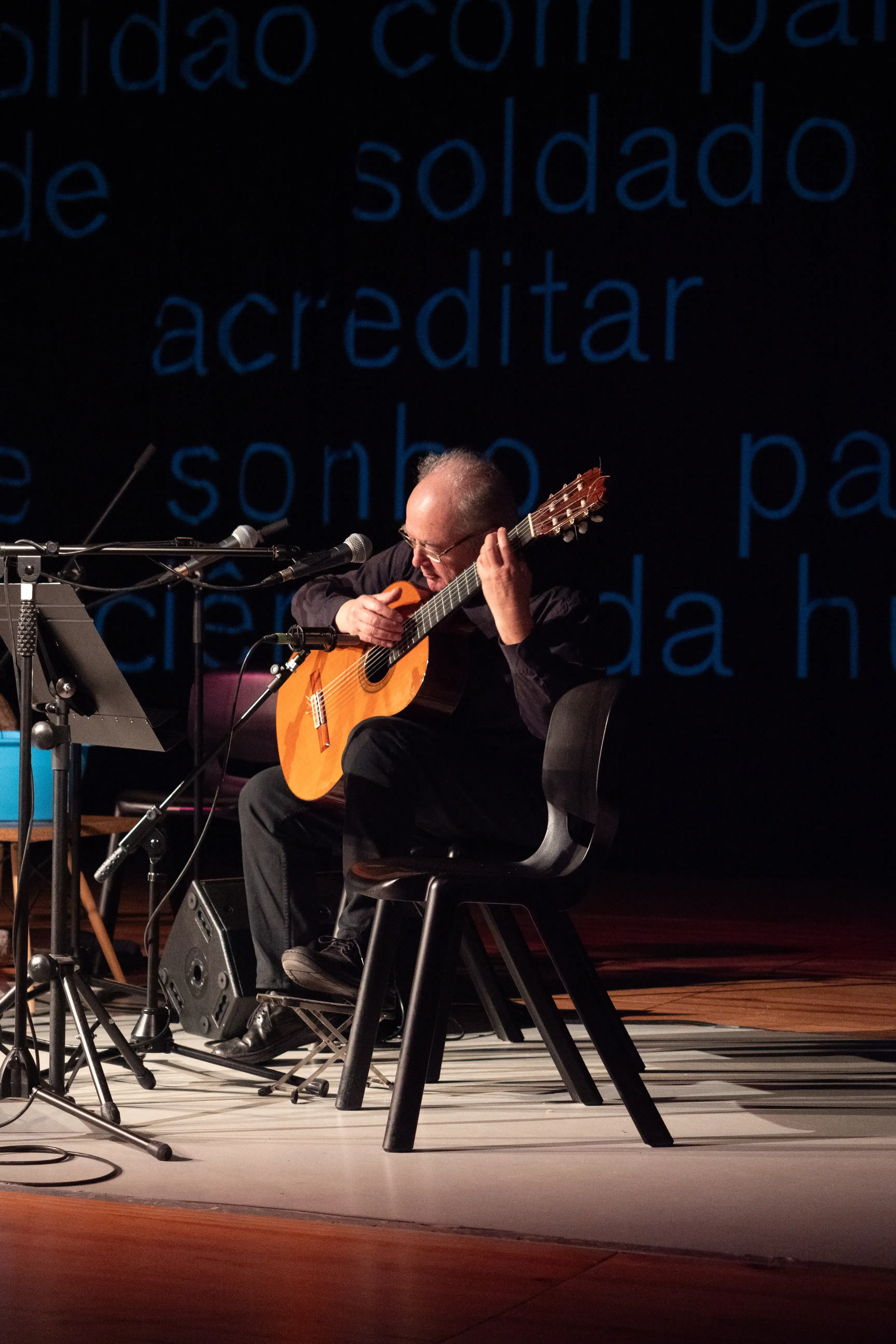 Homem a tocar guitarra clássica. Ao fundo estão projetadas palavras dispersas azuis.